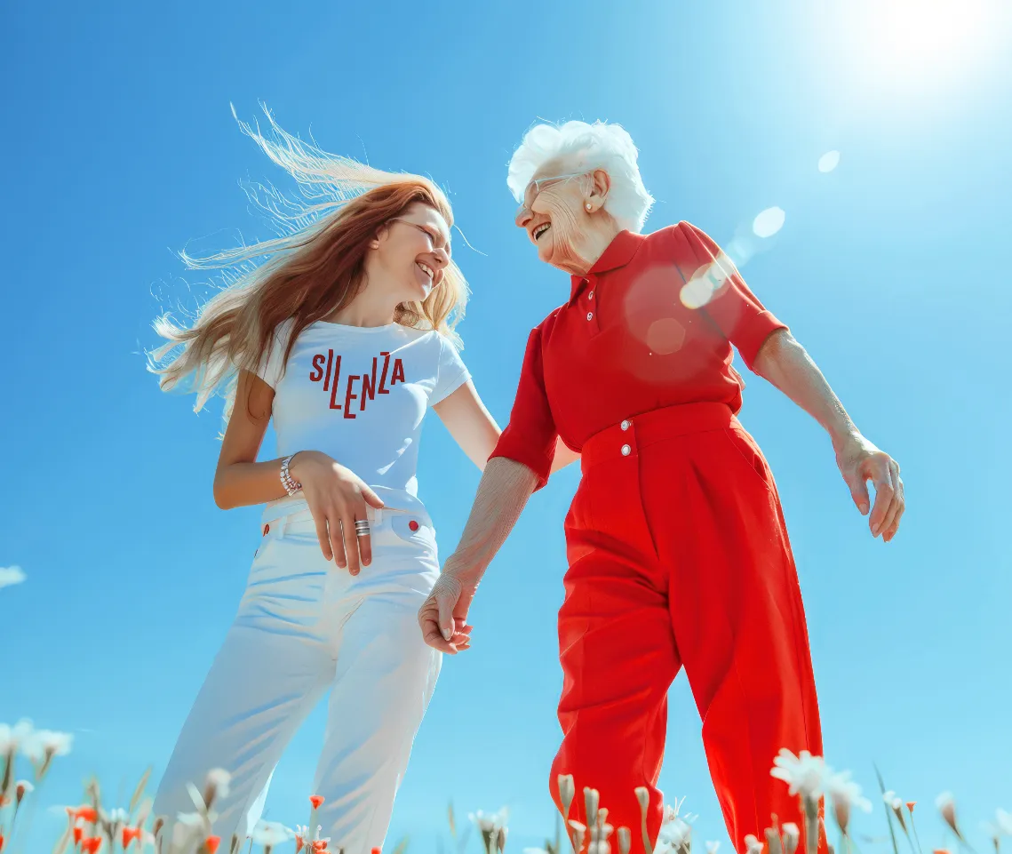 Junge Frau im Silenza-Shirt und lachende Seniorin gehen Hand in Hand über eine Blumenwiese