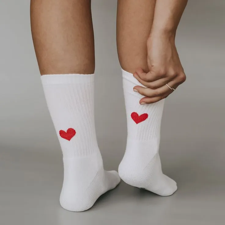 Beine in weißen Sportsocken mit aufgesticktem rotem Herz, eine Hand zieht eine Socke zurecht